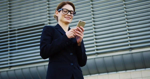 Портрет белой красавицы в очках и черной куртке, заклеенной на смартфон, улыбающейся и смотрящей в сторону возле большого офисного центра. На открытом воздухе — стоковое фото