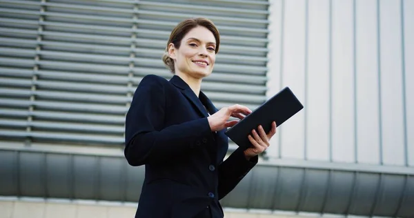 Портрет белой красивой женщины в очках и черной куртке, заклеенной на планшетный компьютер и улыбающейся в камеру возле большого офисного здания. На открытом воздухе — стоковое фото