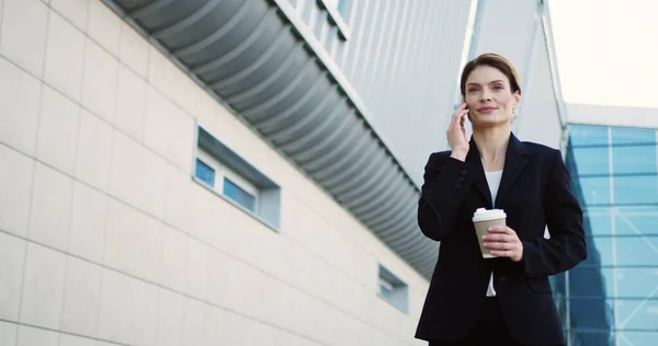 Attraktiv leende affärskvinna promenader på glas kontor centrum bakgrund, prata i telefon och hålla sin morgon kaffe på en solig dag. Utomhus — Stockfoto