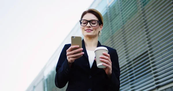 Белая красивая деловая леди в черной куртке и очках, заклеивающая и прокручивающая на смартфон и пьющая утренний кофе. Большое офисное здание позади. На открытом воздухе — стоковое фото