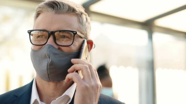 Close up of joyful positive adult senior main glasses wearing mask speaking on smartphone while standing in town on street. Happy handsome male calling on mobile phone in good mood outdoors
