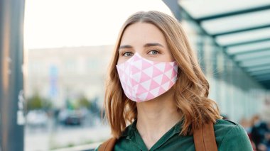 Close up portrait of beautiful Caucasian young woman in face mask standing outdoors on sunny street at bus stop and looking at camera in good mood. Coronavirus pandemic, covid-19 concept