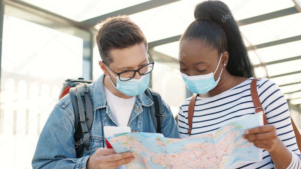 Close up portrait of multi-ethnic young couple man and woman travelling in medical masks during covid pandemic standing outdoors and looking at city map searching for direction, travel concept