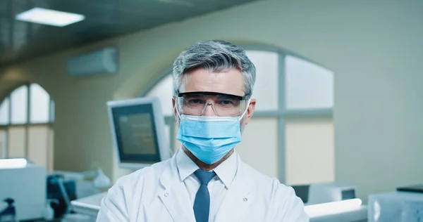 Close up retrato de alegre positivo caucasiano barbudo médico profissional masculino em máscara médica em pé no laboratório de clínica olhando para câmera e sorrindo. Laboratório hospitalar, pandemia de coronavírus — Fotografia de Stock