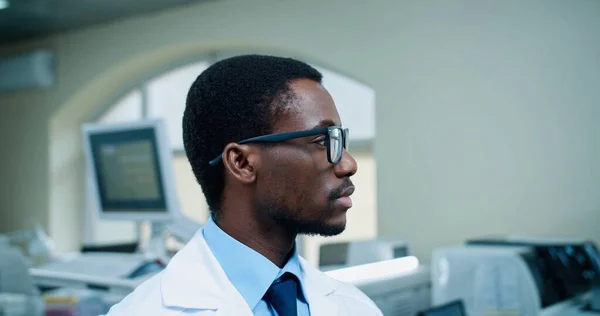 Nahaufnahme eines lebenslustigen jungen, gut aussehenden afroamerikanischen Arztes mit Brille, der in die Kamera blickt und am Arbeitsplatz im Krankenhauslabor lächelt. Medizinische Fachangestellte, Laborfachkraft, Medizinkonzept — Stockfoto