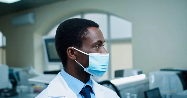 Schöner afroamerikanischer junger Mann mit medizinischer Maske blickt gut gelaunt in die Kamera am Arbeitsplatz im Krankenhauslabor. Männlicher Arzt im klinischen Labor, Coronavirus. Nahaufnahme — Stockfoto