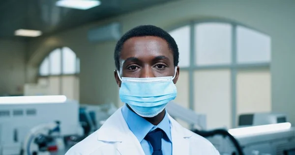 Schöner afroamerikanischer junger Mann mit medizinischer Maske blickt gut gelaunt in die Kamera am Arbeitsplatz im Krankenhauslabor. Männlicher Arzt im klinischen Labor, Coronavirus. Nahaufnahme — Stockfoto