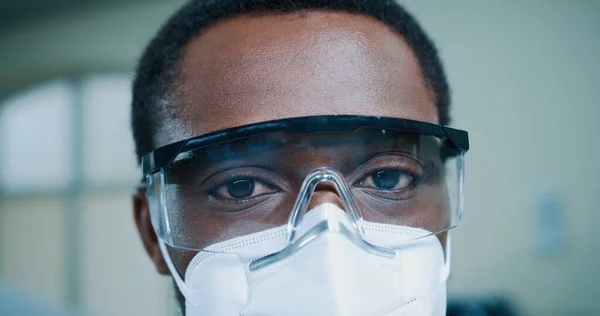 Primo piano del giovane medico afro-americano mascherato e con gli occhiali che guarda la telecamera al lavoro in clinica. Uomo specializzato in laboratorio, ospedale, concetto di medicina, pandemia di coronavirus, operaio medico — Foto Stock
