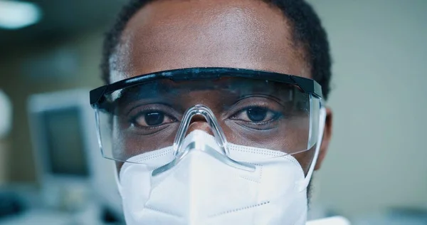 Primer plano de joven afroamericano médico en máscara y gafas mirando a la cámara en el trabajo en la clínica. Hombre especialista en laboratorio, hospital, concepto de medicina, pandemia de coronavirus, trabajador médico —  Fotos de Stock