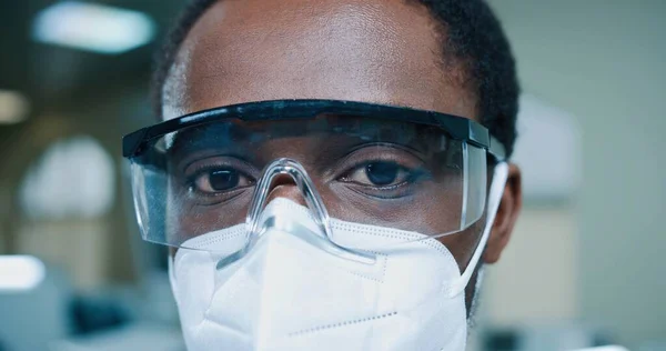 Close up de jovem afro-americano médico masculino em máscara e óculos olhando para câmera no trabalho na clínica. Homem especialista em laboratório, hospital, medicina conceito, coronavírus pandêmico, médico trabalhador — Fotografia de Stock