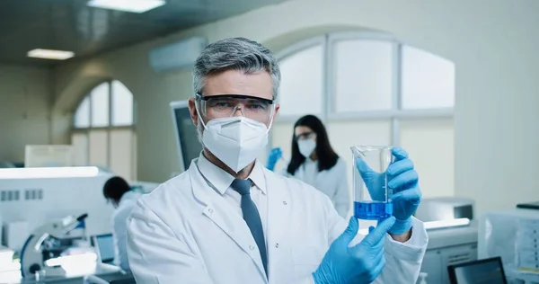 실험실에서 파란색 액체가 들어간 제품을 카메라를 코카서스 약사의 과학자가고 마스크를 — 스톡 사진