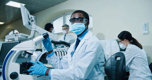 Retrato de jovem experiente médico afro-americano especialista sentado à mesa no laboratório do hospital trabalhando em equipamentos modernos de microscópio olhando para a análise. Cientista médico, conceito de experiência — Fotografia de Stock