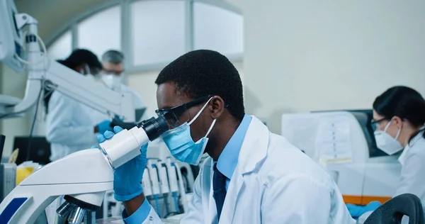 Nahaufnahme eines jungen ernstzunehmenden afroamerikanischen Mannes mit Maske, der am Schreibtisch im klinischen Labor sitzt, an modernen Mikroskopgeräten arbeitet und in die Kamera blickt. Medizinischer Wissenschaftler, lebendige Behandlung — Stockfoto