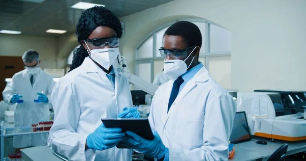 Retrato de afro-americanos jovens especialistas médicos masculinos e femininos em máscaras e casacos brancos em pé no laboratório de clínica e tocando no tablet discutindo resultados de análise. Saúde, conceito de laboratório — Fotografia de Stock