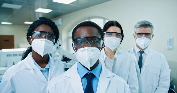 Close up of mixed-race different doctors in masks and goggles standing in clinic lab and looking at camera. Team of male and female medical scientists at lab. Healthcare, medicine, science concept — Zdjęcie stockowe