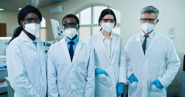 Retrato de mestiços diferentes especialistas médicos em máscaras e óculos em pé no laboratório do hospital e olhando para a câmera. Equipa de cientistas masculinos e femininos no laboratório. Saúde, medicina, ciência — Fotografia de Stock