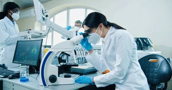 Visão lateral da jovem especialista médica branca em máscara e luvas sentadas em laboratório usando microscópio moderno procurando resultados de análise. Medicina, pesquisador médico no laboratório — Fotografia de Stock