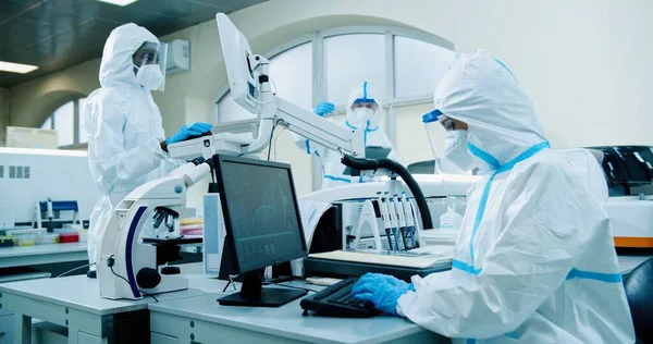 Mixed-race medical scientists wearing protective uniforms, masks and face shields working in modern laboratory during coronavirus typing on computer and medical equipment. Clinical research concept