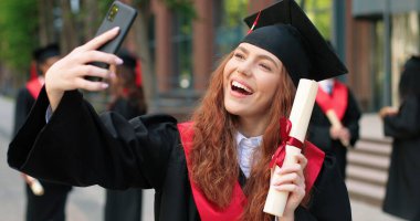 Selfie with diploma. Happy caucasian student girl being happy at her graduation day. Attractive woman looking at the smartphone and making selfie