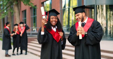 Two happy multiracial student people are celebrating their graduation while standing near the college and rejoicing with diplomas at the hands. Education concept