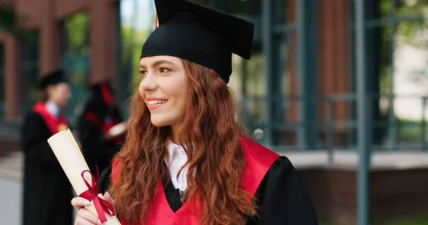 School graduate in academic dress and hat looking at the camera with happy smile. Adorable girl rejoicing near her university or school. Graduation concept