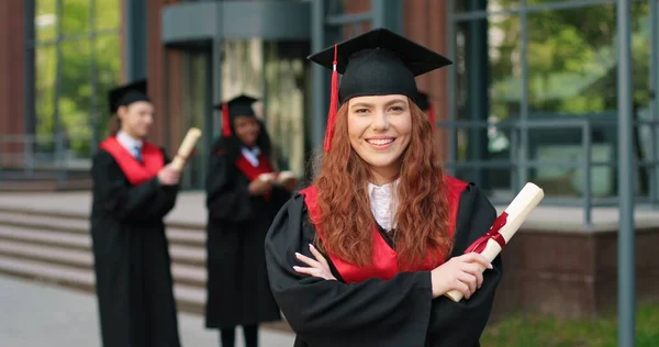 Schulabsolventen in akademischer Kleidung und mit Hut blicken mit glücklichem Lächeln in die Kamera. Entzückendes Mädchen, das in der Nähe ihrer Universität oder Schule jubelt. Graduierungskonzept — Stockfoto