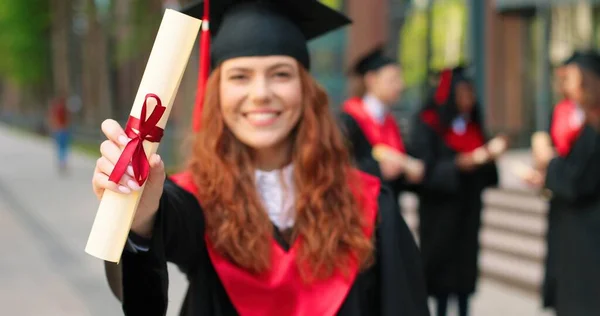 Pädagogisches Konzept. Glückliche kaukasische Studentin, die glücklich ist über ihr Diplom, ihren Abschluss. Ingwerfrau steht mit ihren Freunden im Hintergrund auf der Straße — Stockfoto