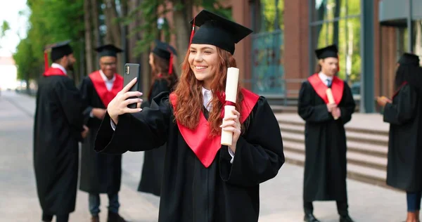Selfie mit Diplom. Glückliches kaukasisches Studentenmädchen, das an ihrem Abi-Tag glücklich ist. Attraktive Frau schaut aufs Smartphone und macht Selfie — Stockfoto