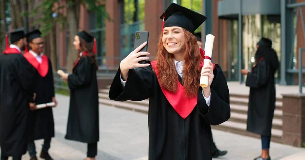 有文凭的自私自利。快乐的高加索学生女孩是快乐的在她的毕业典礼上。迷人的女人看着智能手机，自私自利 — 图库照片