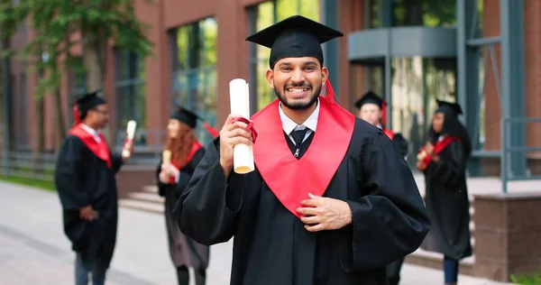 Schulabsolvent in akademischem Gewand und Hut, der mit glücklichem Lächeln in die Kamera blickt. Glücklicher multiethnischer Mann, der in der Nähe seiner Universität oder Schule jubelt. Graduierungskonzept — Stockfoto