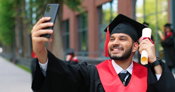 Selfie com diploma. Menino estudante multirracial feliz sendo feliz em seu dia de formatura. Homem animado olhando para o smartphone e fazendo selfie com diploma — Fotografia de Stock