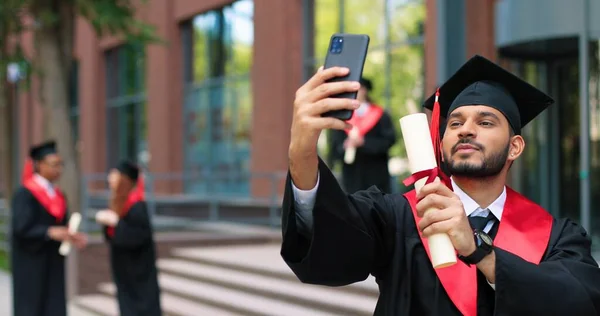 有文凭的自私自利。快乐的多种族学生男孩在他的毕业典礼上很快乐。兴奋的男人看着智能手机，拿着文凭自立门户 — 图库照片