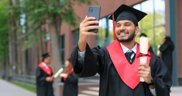 Selfie com diploma. Menino estudante multirracial feliz sendo feliz em seu dia de formatura. Homem animado olhando para o smartphone e fazendo selfie com diploma — Fotografia de Stock