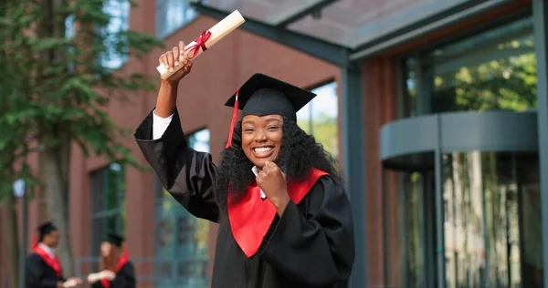 School graduate woman in academic gown and hat looking at the camera with happy smile. Happy multiracial girl rejoicing near her university or school. Graduation concept — Photo