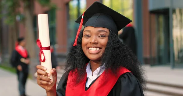 School graduate woman in academic gown and hat looking at the camera with happy smile. Happy multiracial girl rejoicing near her university or school. Graduation concept — Stock Photo, Image