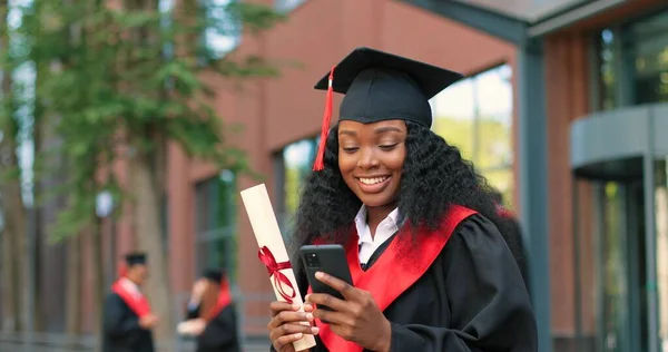 E-learning, education and people concept. Happy smiling multiracial female graduate student standing at the street with smartphone and typing message with pleasure smile — Photo