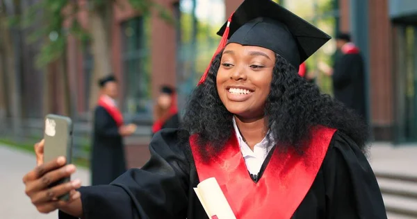 Concepto de aprendizaje electrónico, educación y personas. Feliz estudiante graduada multirracial sonriente de pie en la calle con teléfono inteligente y escribiendo mensaje con sonrisa de placer —  Fotos de Stock