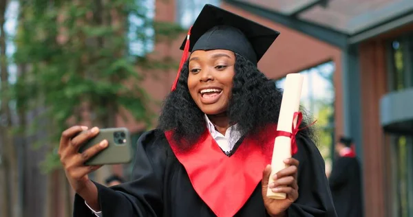 Portrait of a happy attractive multiracial graduate holding her smartphone and chatting via video call. College university graduation day. Proud woman towards camera with big smile — Photo
