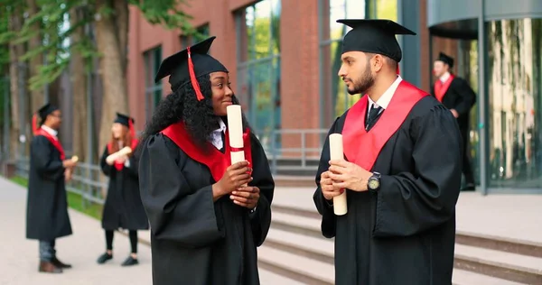 Two happy multiracial student people are celebrating their graduation while standing near the college and rejoicing with diplomas at the hands. Education concept — Stock fotografie