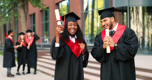 Two happy multiracial student people are celebrating their graduation while standing near the college and rejoicing with diplomas at the hands. Education concept — Stock Fotó