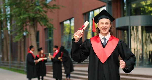 Schulabsolvent in akademischem Gewand und Hut, der mit glücklichem Lächeln in die Kamera blickt. Glücklicher kaukasischer Junge, der in der Nähe seiner Universität oder Schule jubelt. Graduierungskonzept — Stockfoto