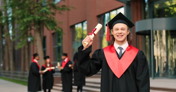 School graduate man in academic gown and hat looking at the camera with happy smile. Happy caucasian boy rejoicing near his university or school. Graduation concept — ストック写真