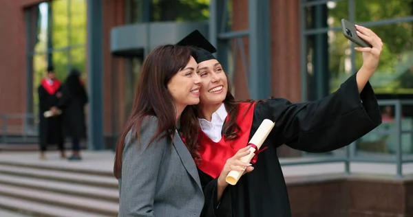 Mãe feliz e sua filha graduada posando para selfie no dia da formatura. Menina em vestido acadêmico e bonés tirando auto foto com sua mãe orgulhosa — Fotografia de Stock