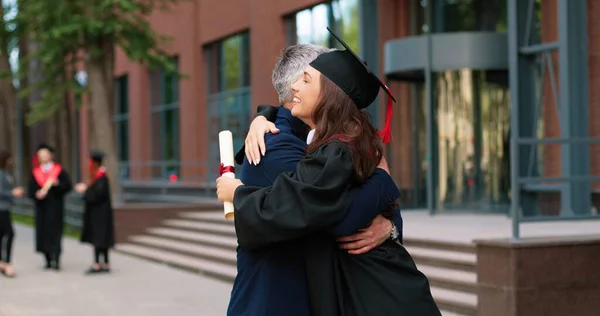 Happy female graduate are embracing with her father with diploma at her hands and rejoicing with each other. Man is hugging his daughter with other students moving and talking in background — ストック写真
