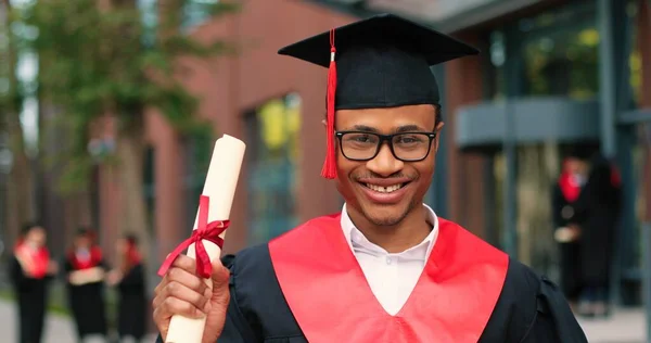 穿着校服戴帽子的毕业生开心地看着摄像机。在他的大学或学校附近快乐的多种族人。毕业概念 — 图库照片
