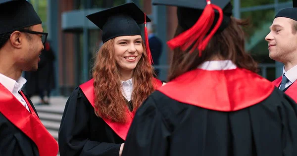 Nahaufnahme der fröhlichen Studenten mit Mützen und Kleidern, die sich untereinander unterhalten, während sie ihren Universitätsabschluss feiern. Bildungskonzept — Stockfoto