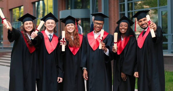 This is final. Full length view of the group of students at the graduation gowns and caps standing near the university and showing their diplomas to the camera