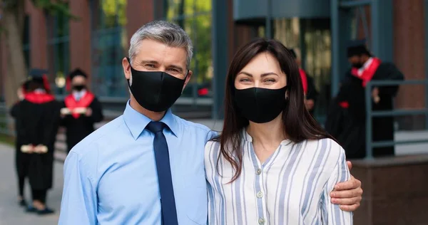 Vista del retrato de los tranquilos padres felices con máscara protectora mirando a la cámara y sonriendo mientras esperan a su hijo después de la graduación. Concepto educativo — Foto de Stock