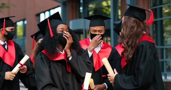 Unforgettable emotions. Waist up portrait of the group of students at the graduation gowns and caps wearing protective masks standing near the university and sharing their emotions with each other — ストック写真