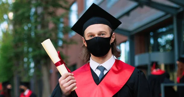 Portrait view of the caucasian male student wearing black hat and protective mask posing to the camera because he graduated from university during the covid 19 pandemic. Education concept — ストック写真